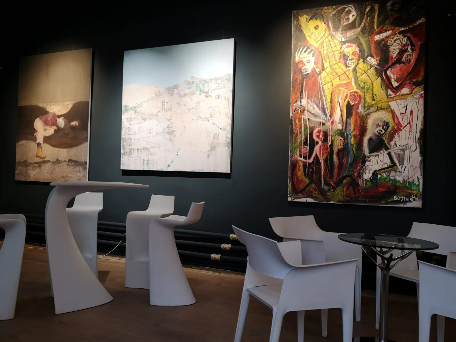 Large original paintings displayed against dark walls beside white sculptural furniture.